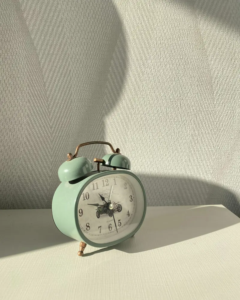 Ein mintgrüner Wecker steht in schrägem Fensterlicht vor einer hellen Wand.